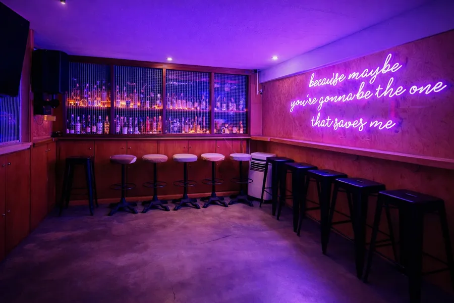 Karaoke Room 3: bar stools along a plywood counter, spirits behind a mesh grille and a neon lyric on the wall
