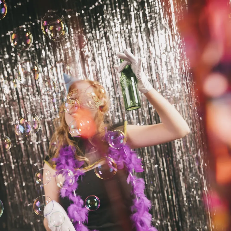 A partygoer in a feather boa and mask blowing bubbles under a sparkling backdrop